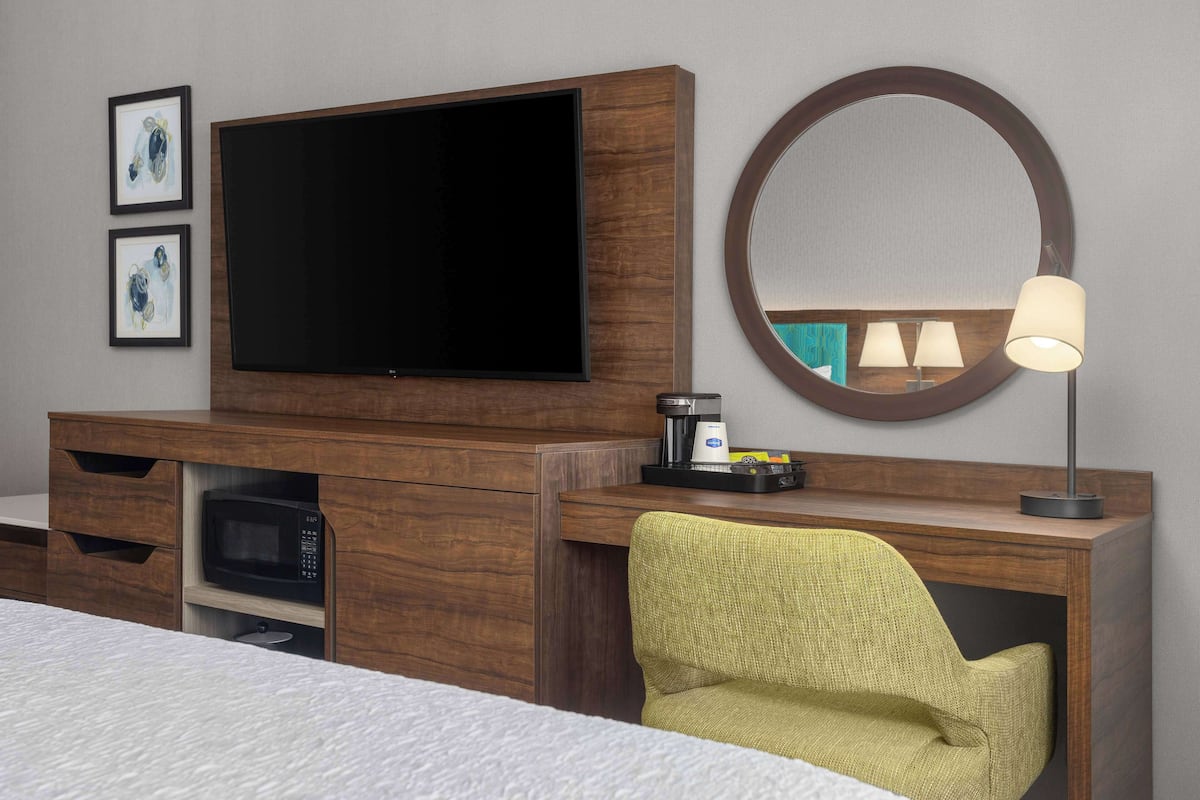 Una habitación de hotel con una cama, un televisor de pantalla plana, una cafetera, un microondas, un sillón verde y un espejo redondo.