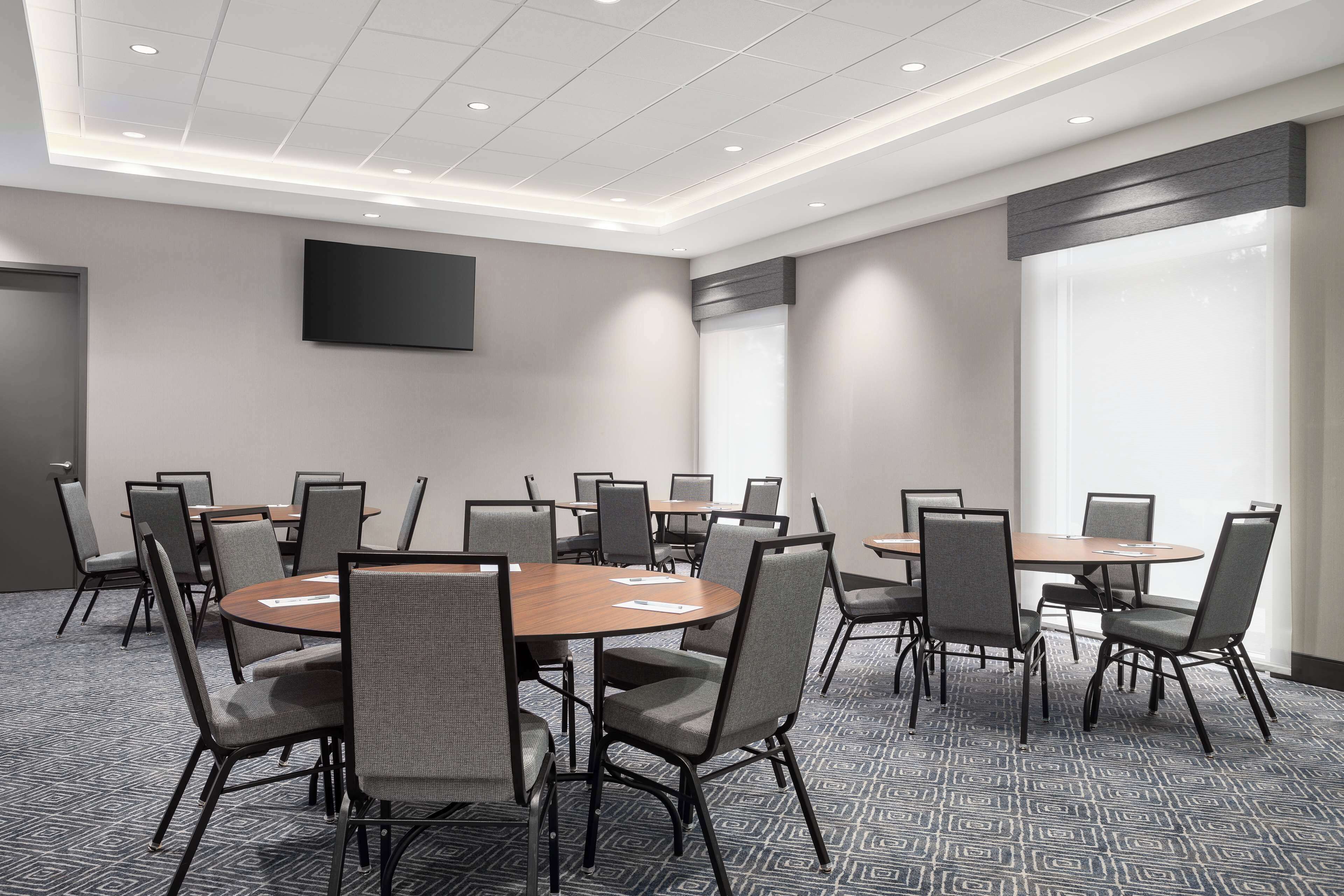 A conference room with round tables and chairs, a flat-screen TV on the wall, and large windows.