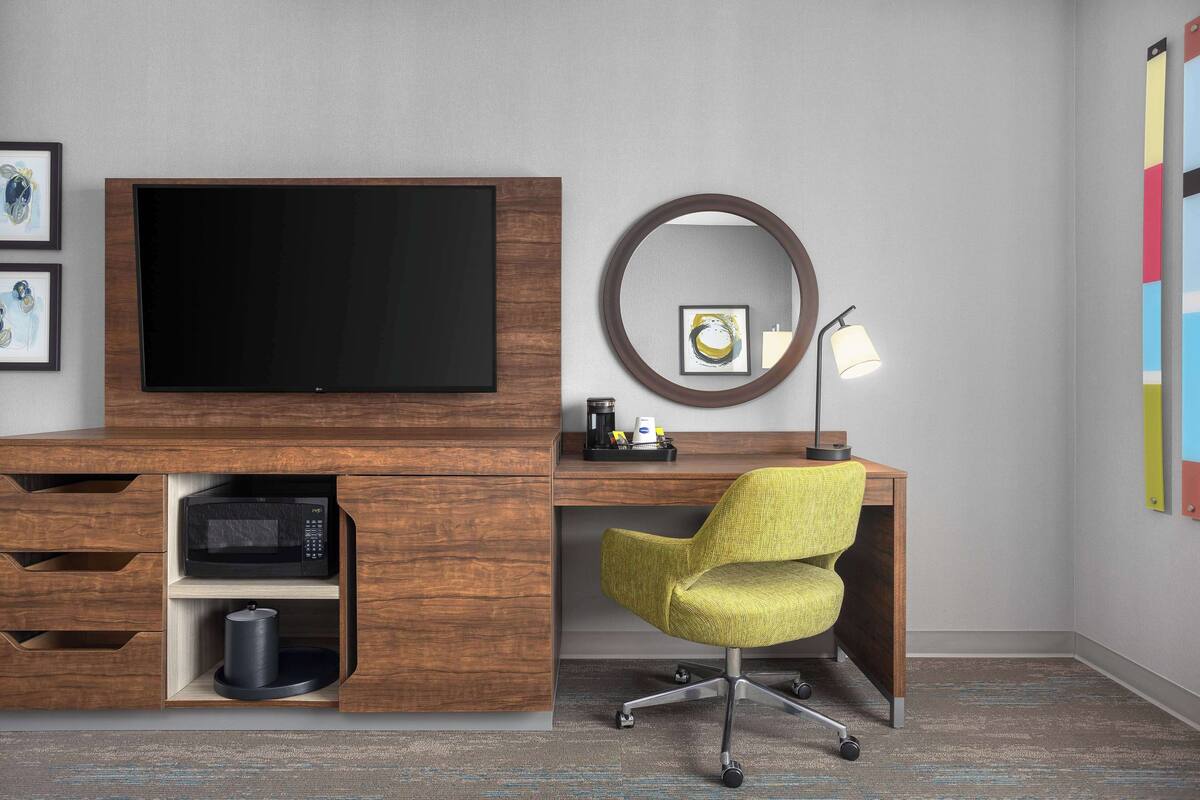 Un espacio de oficina moderno con un escritorio de madera, una silla amarilla, un televisor montado, un espejo redondo y una lámpara.