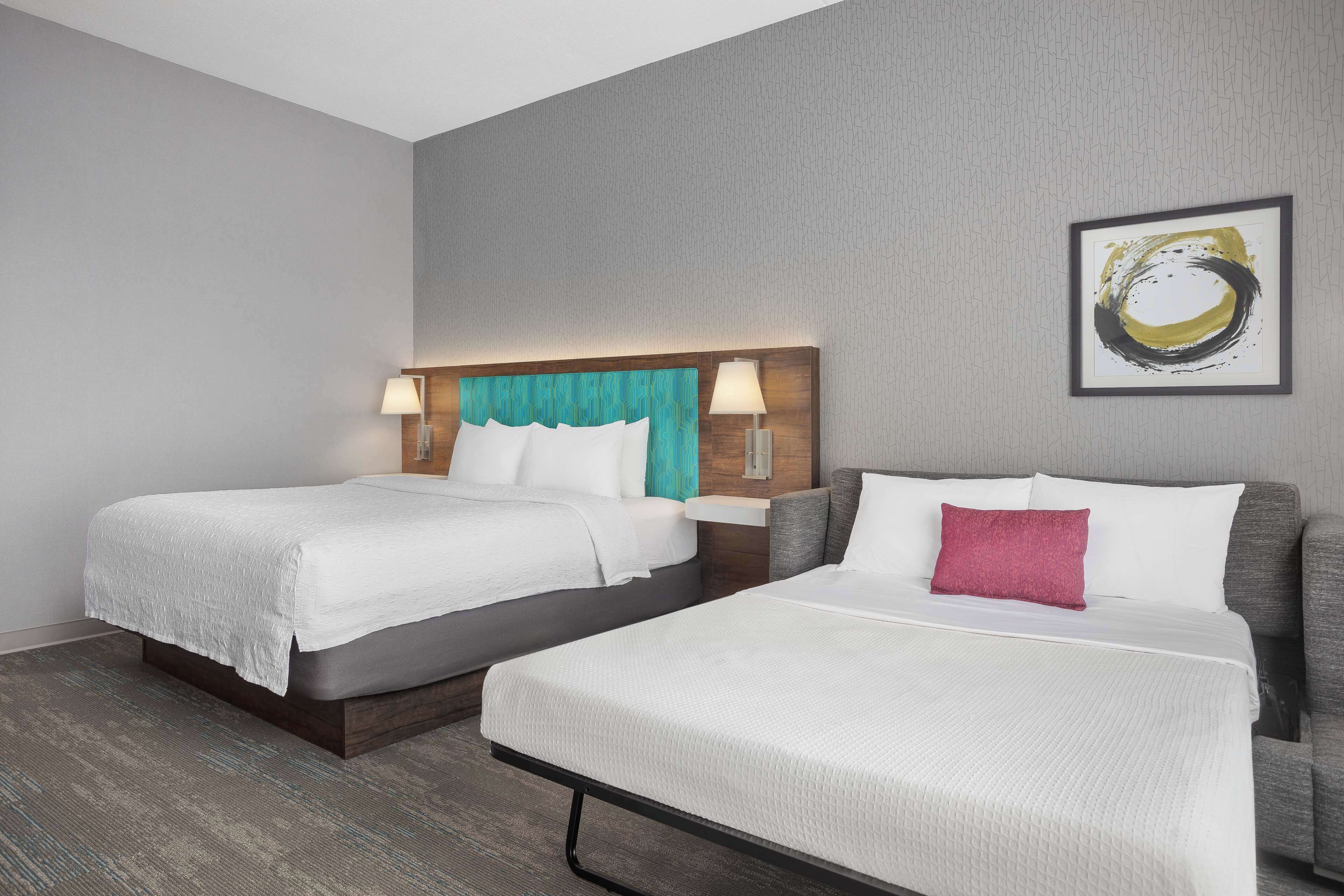 Quarto de hotel com duas camas, parede cinza texturizada, pintura abstrata emoldurada e cabeceira de madeira.