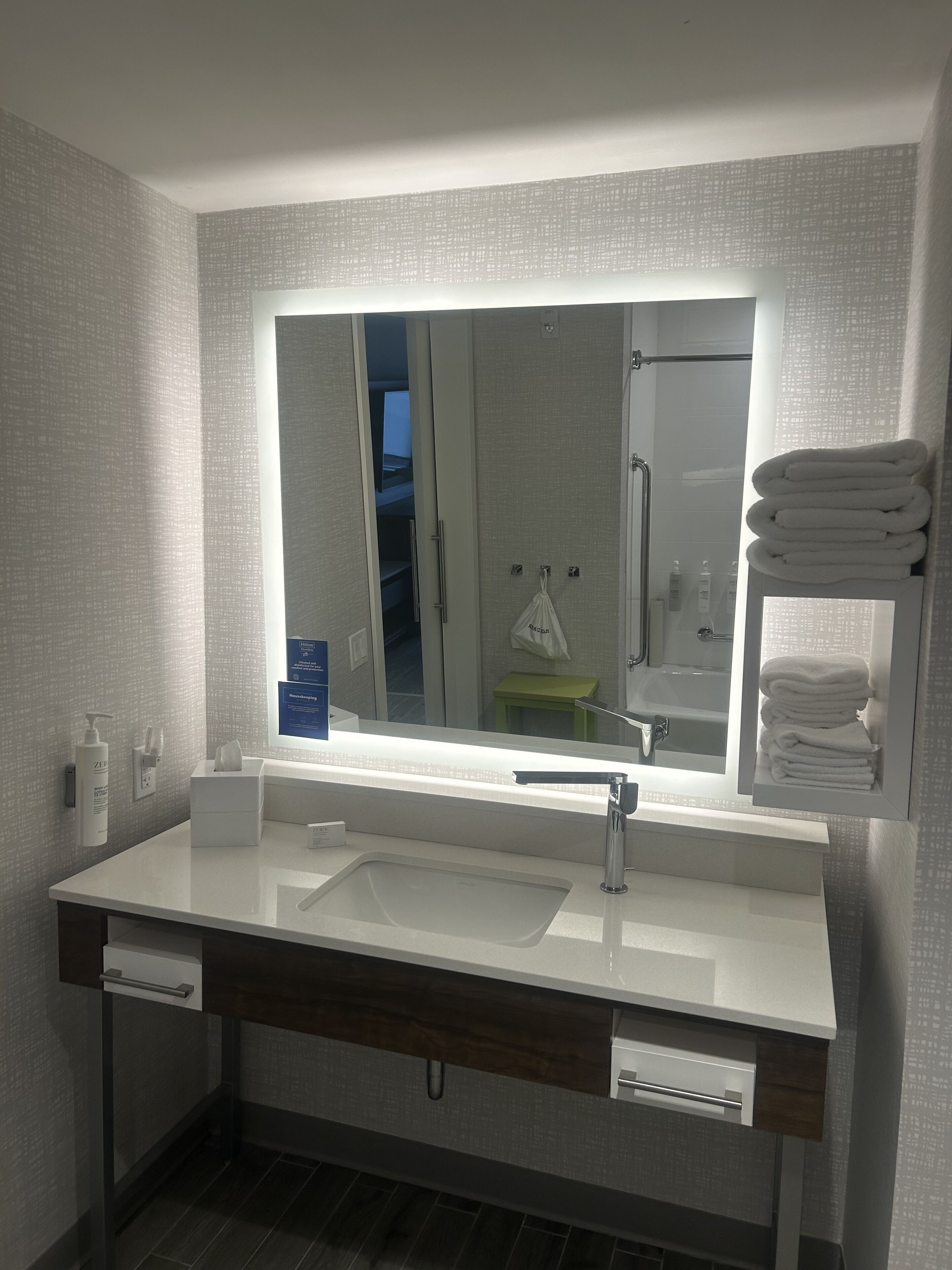 A modern bathroom with a large mirror, two sinks, a towel rack, and a toilet.
