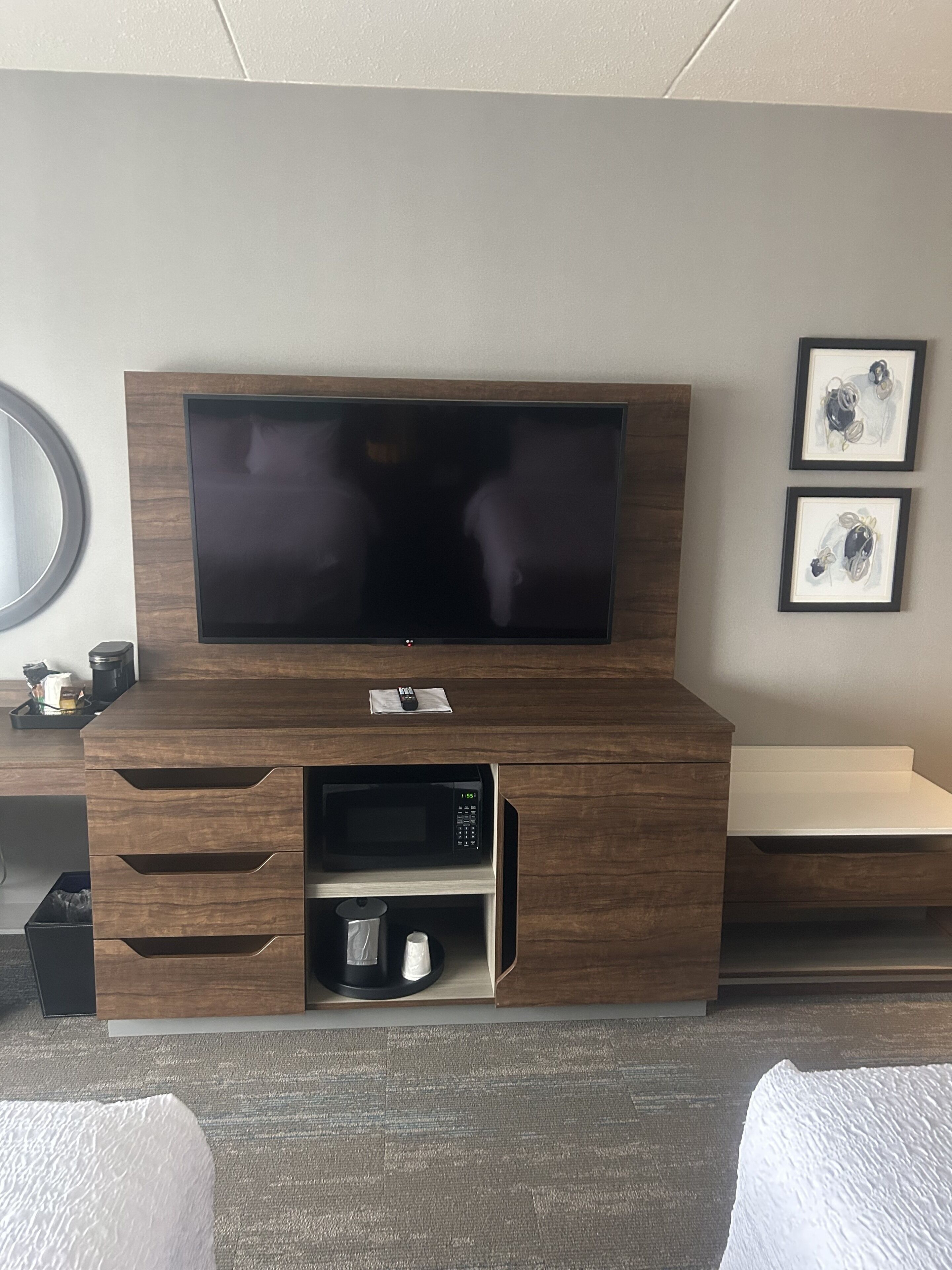 A hotel room with a large flat-screen TV mounted on a wooden cabinet, a microwave, and a small shelf below. There are two framed pictures on the wall and a round mirror on the left.