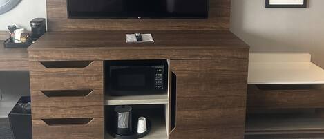 A hotel room with a large flat-screen TV mounted on a wooden cabinet, a microwave, and a small shelf below. There are two framed pictures on the wall and a round mirror on the left.