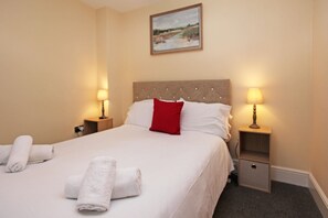 A neatly made bed with white linens and a red pillow, two bedside lamps, and a small drawer under the bed. - St Julians Place (Shrewsbury)