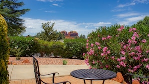 A patio with a round metal table and chairs, surrounded by lush greenery and pink flowers, with a view of red rock formations in the distance.
