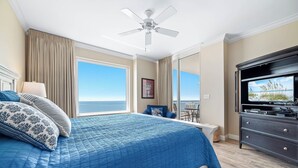 A bedroom with a large bed, a ceiling fan, a TV, and a view of the ocean.