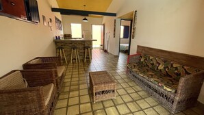 A spacious room with a tiled floor, a wooden sofa with a floral pattern, wicker furniture, and a kitchen area with stools.