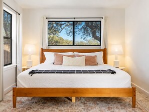 A neatly made bed with a wooden frame, white bedding, and a view of trees through a window.