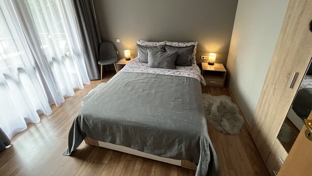 A modern bedroom with a large bed, two bedside tables with lamps, a chair, and a view through sheer curtains. - MH Apartaments (Bansko)