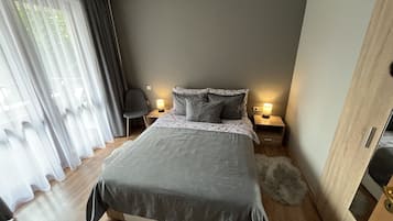 Une chambre moderne avec un grand lit, deux tables de chevet avec des lampes, un fauteuil et une vue à travers des rideaux légers.