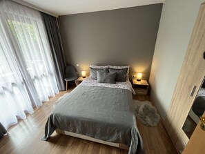 Une chambre moderne avec un grand lit, deux tables de chevet avec des lampes, un fauteuil et une vue à travers des rideaux légers.