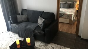 A modern living room with a dark grey sofa, two side tables with candles, a marble-patterned rug, and a view into a bedroom with a bed and a nightstand. - MH Apartaments (Bansko)