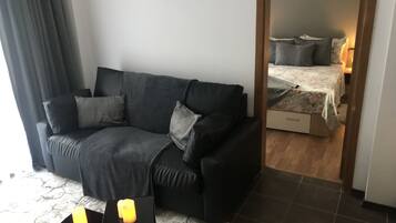 Un salon moderne avec un canapé gris foncé, deux tables d’appoint ornées de bougies, un tapis à motifs de marbre et une vue sur une chambre à coucher avec un lit et une table de chevet.