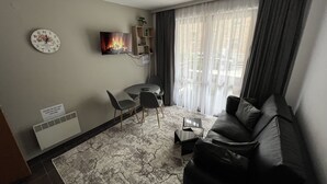 A living room with a sofa, a TV, a clock, a small table, and a chair. - MH Apartaments (Bansko)