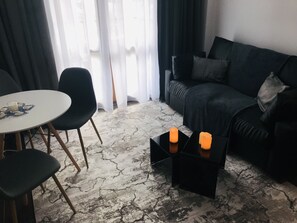 A modern living room with a black leather sofa, a round dining table with chairs, and a coffee table. There are two candles on a black side table.