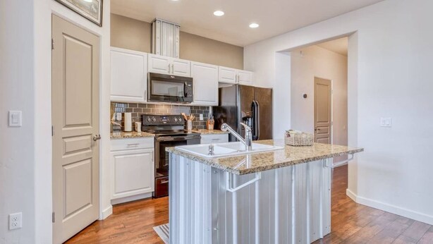 Una cocina moderna con armarios blancos, una isla con encimera de granito, electrodomésticos de acero inoxidable y suelo de madera.
