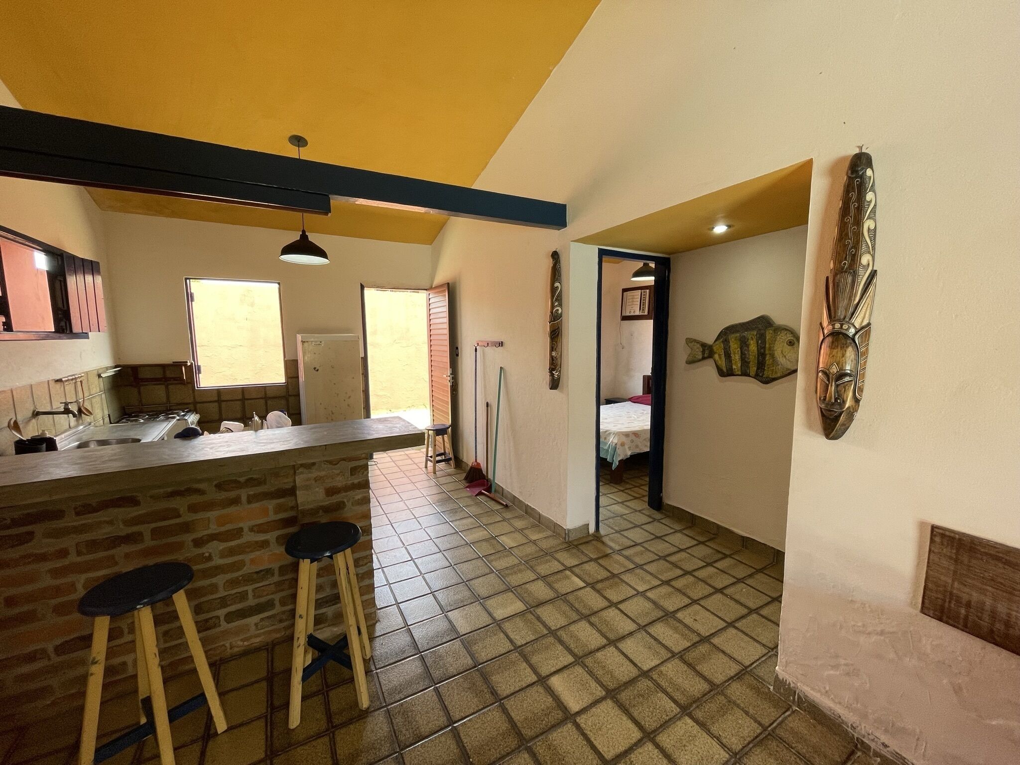 A kitchen with a central island, stools, a tiled floor, and a view into a bedroom.