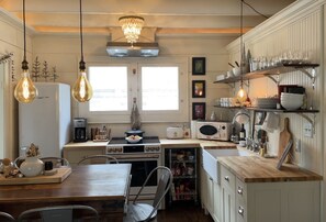 Een keuken met een houten tafel, twee hangende lampen, een kroonluchter, een koelkast, een fornuis, en planken met servies en glazen.
