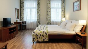 A hotel room with a bed, bedside tables, a desk with a chair, a television, and curtains. - Orion (Prague)