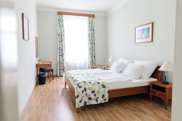 Habitación de hotel ordenada con una cama de madera, una mesita de noche con lámpara, un escritorio con silla y una ventana con cortinas.
