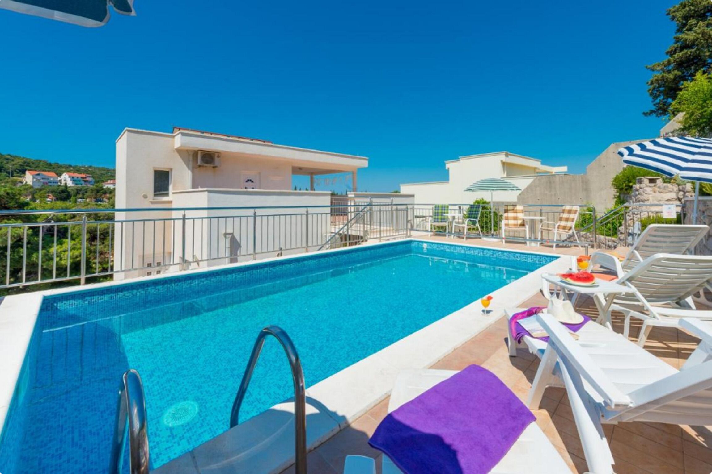 A swimming pool with lounge chairs and umbrellas, surrounded by buildings and a clear sky.