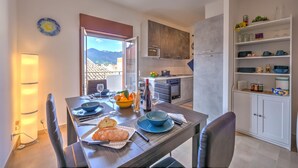 A dining area with a table set for two, a kitchen in the background, and a view of mountains through a window. - Sicilian Charm Close to Taormina (Gaggi)