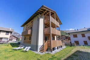 A multi-story building with a wooden balcony, a staircase, and a grassy area with sun loungers.