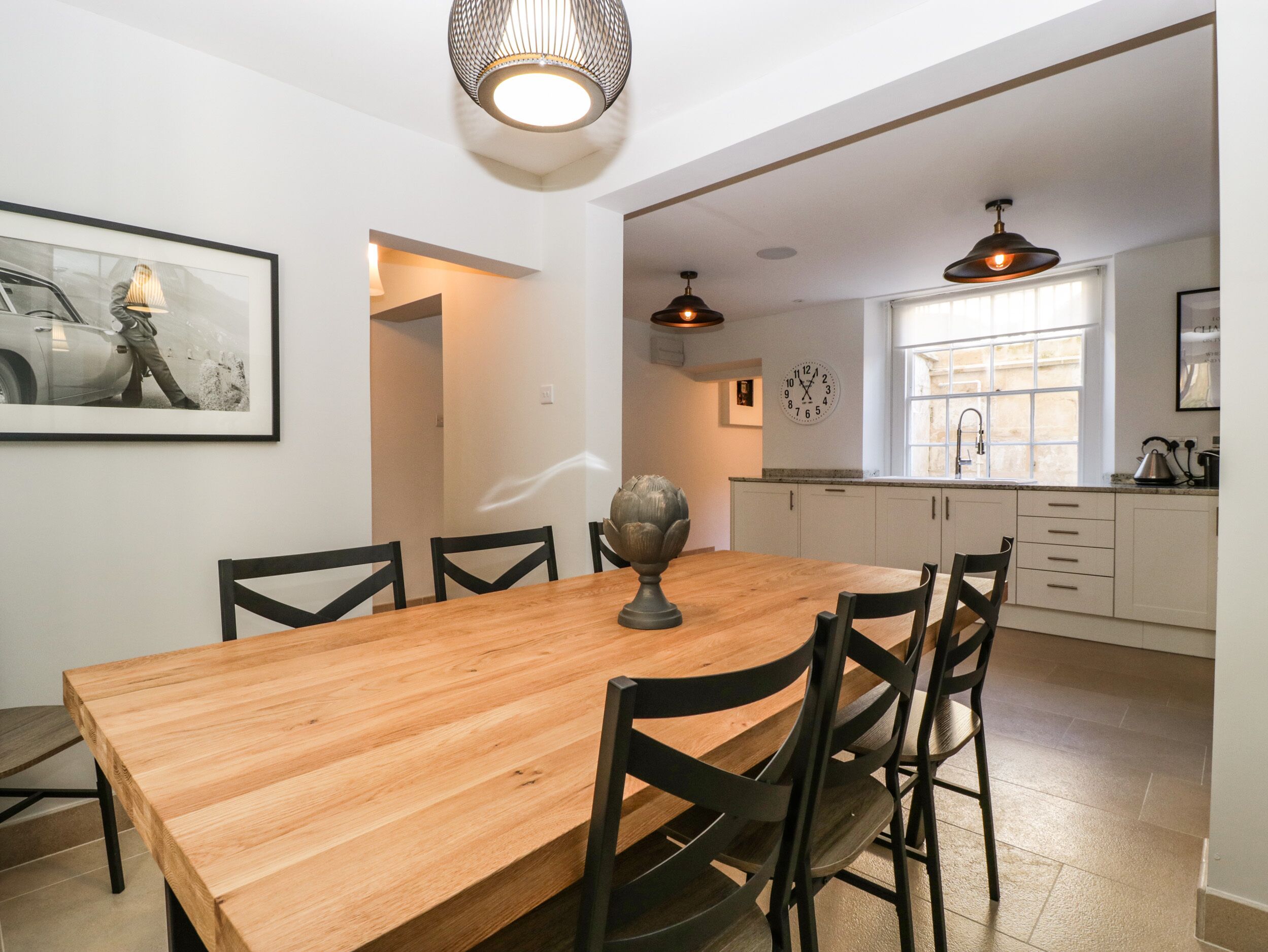 A dining area with a wooden table and chairs, a wall clock, and a kitchen in the background.