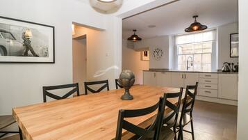 A dining area with a wooden table and chairs, a wall clock, and a kitchen in the background.