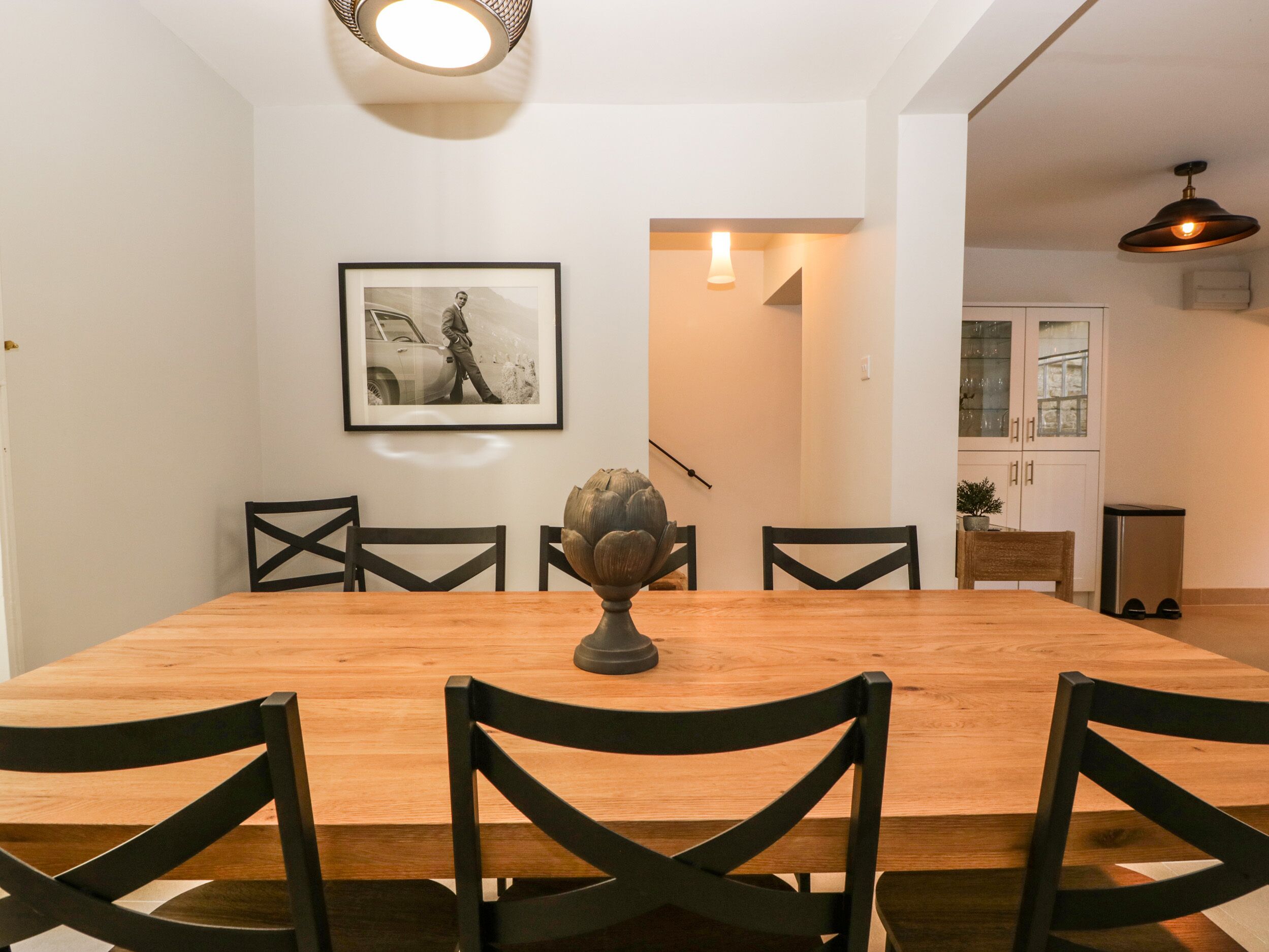 A dining area with a wooden table, black chairs, a framed photograph on the wall, and a staircase leading to another room.