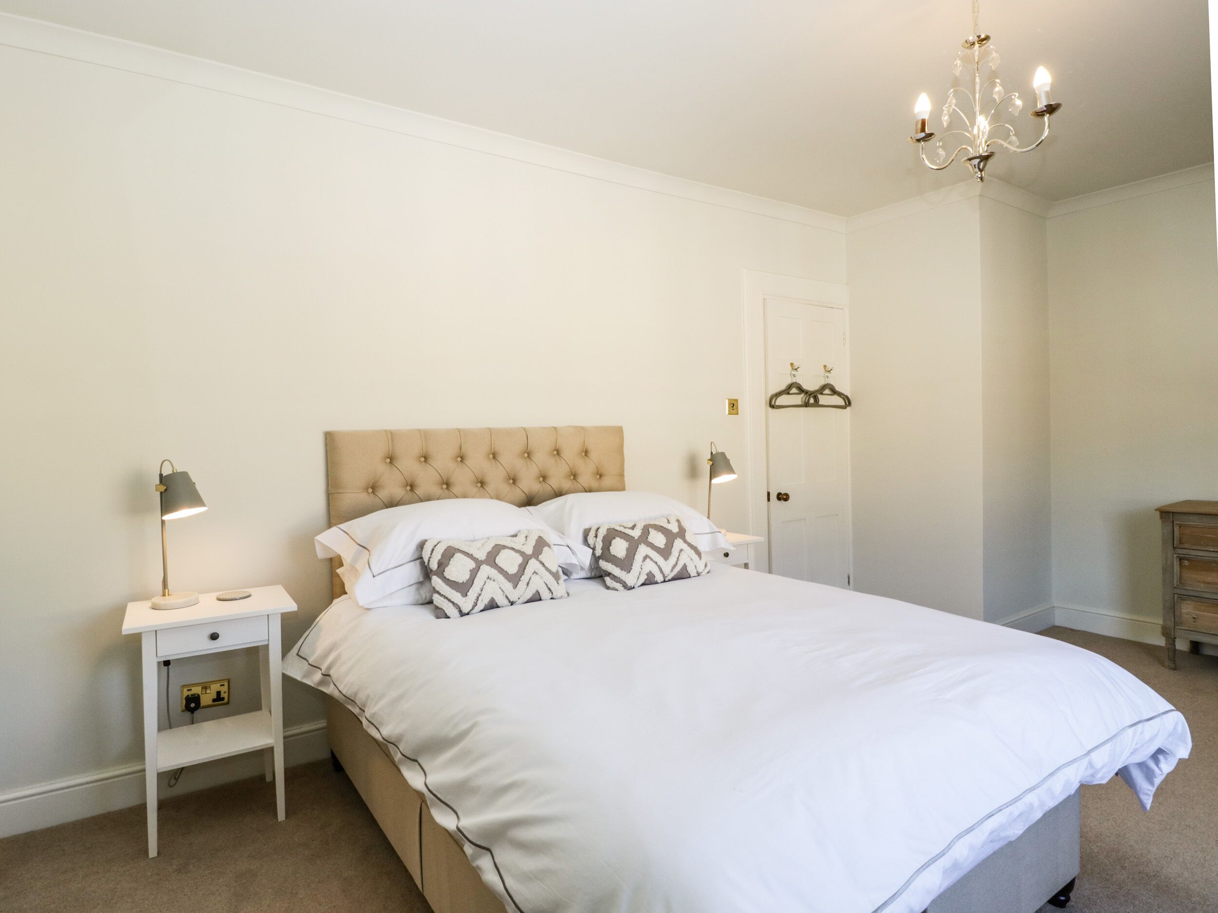 A neatly arranged bedroom with a tufted headboard, a bedside table with a lamp, and a hanging coat rack.