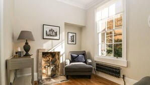 Een kamer met een open haard, een grijze fauteuil, een lamp en een raam met uitzicht op gebouwen.