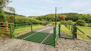 Una entrada rural con un camino de grava que lleva a un portón, rodeada de vegetación y colinas.