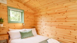 Un dormitorio de cabaña de madera con cama, almohadas, mesita de noche, ventana y una lámpara fijada a la pared.
