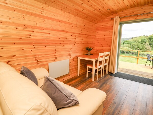 A wooden cabin interior with a sofa, a small table with chairs, and a view of a green landscape through a glass door.