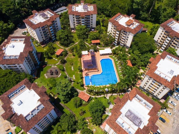 An aerial view of a residential complex with multiple apartment buildings, a central swimming pool, and surrounding greenery.