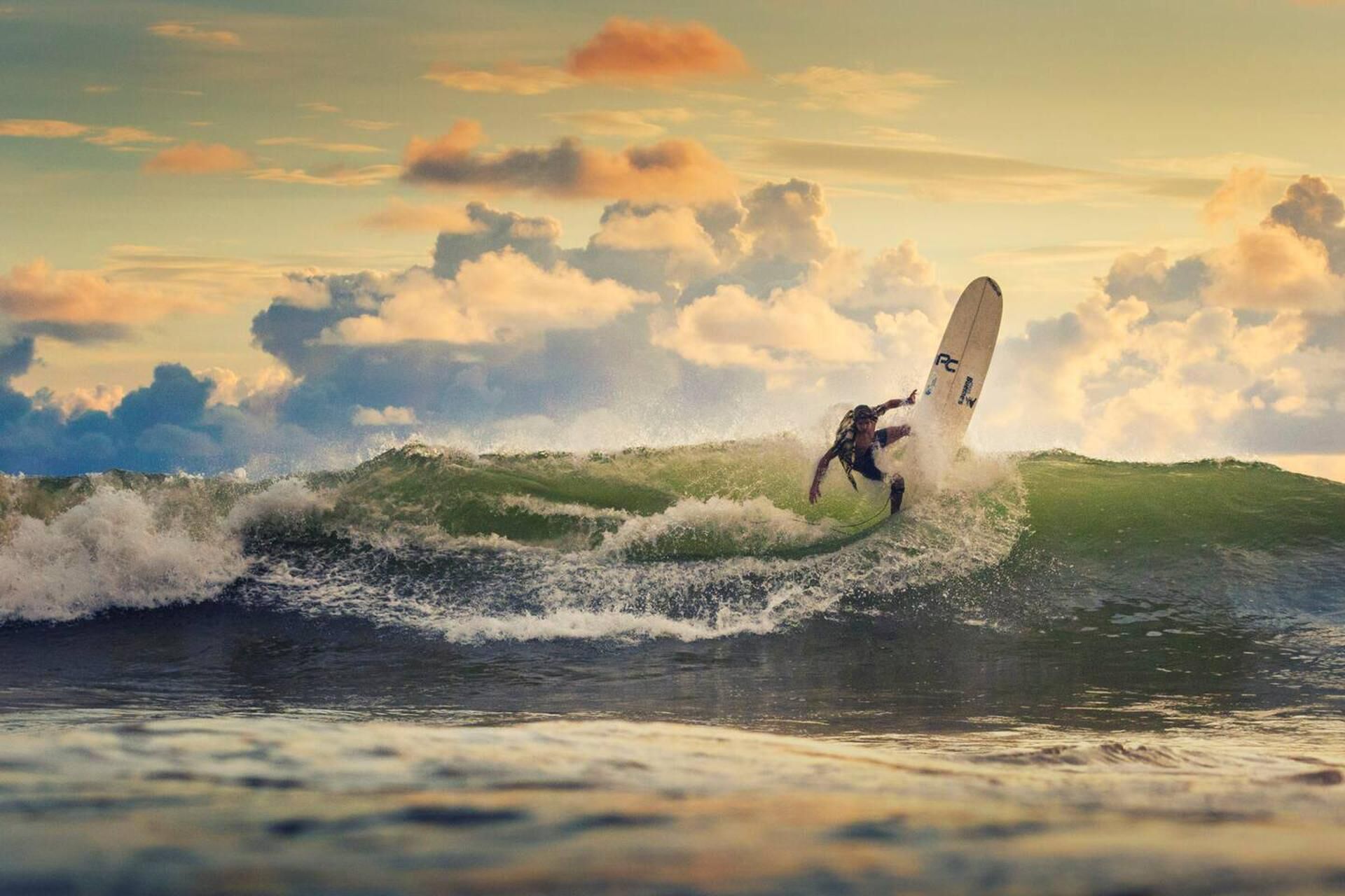 A surfer riding a wave with a surfboard labeled "RC" against a backdrop of a partly cloudy sky.