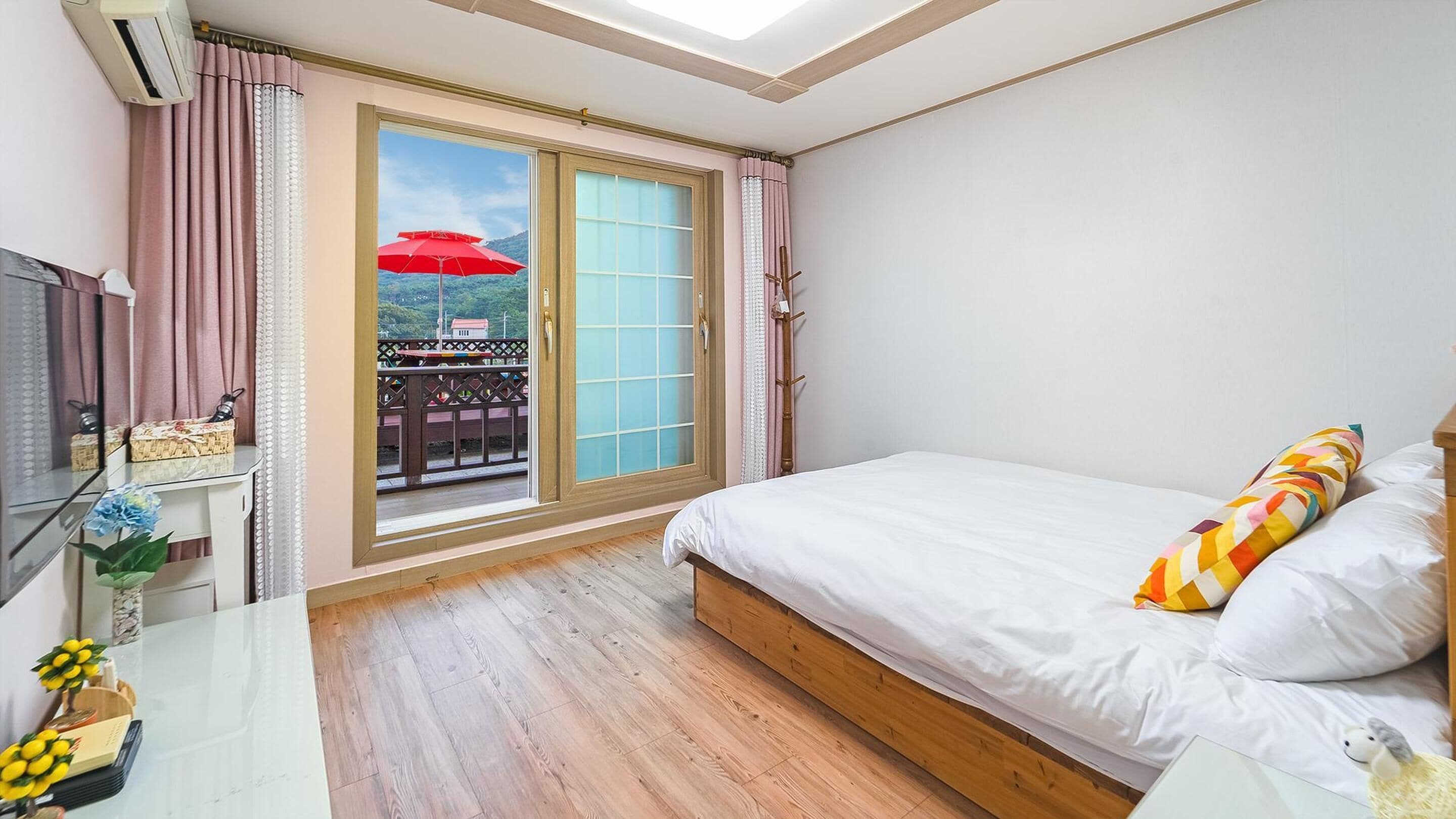 A bedroom with a large sliding glass door leading to a balcony, a bed with white bedding, and a small bedside table with a lamp.