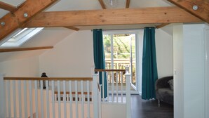 Ein Zimmer mit schräg verlaufender Decke, Holzträgern, einer weißen Geländer und Blick durch eine Glastür auf eine Außenterrasse, sowie ein braunes Sofa.