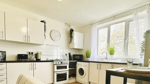A modern kitchen with white cabinets, a stove, a microwave, and a washing machine. There is a window with plants on the windowsill and a clock on the wall. - Beautiful 3-bed Apartment With Driveway Parking (Nottingham)