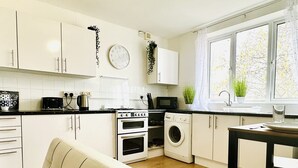 A modern kitchen with white cabinets, a stove, oven, microwave, and a washing machine. There is a window with plants on the windowsill and a dining area with a table and chairs. - Beautiful 3-bed Apartment With Driveway Parking (Nottingham)
