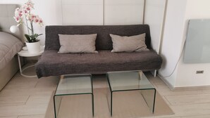A modern living room with a grey sofa, two cushions, a glass coffee table, a small white side table with a potted plant, and a lamp. - Apartment - Sleeps 2 - Parking - 5min To La Frasca (Civitavecchia)