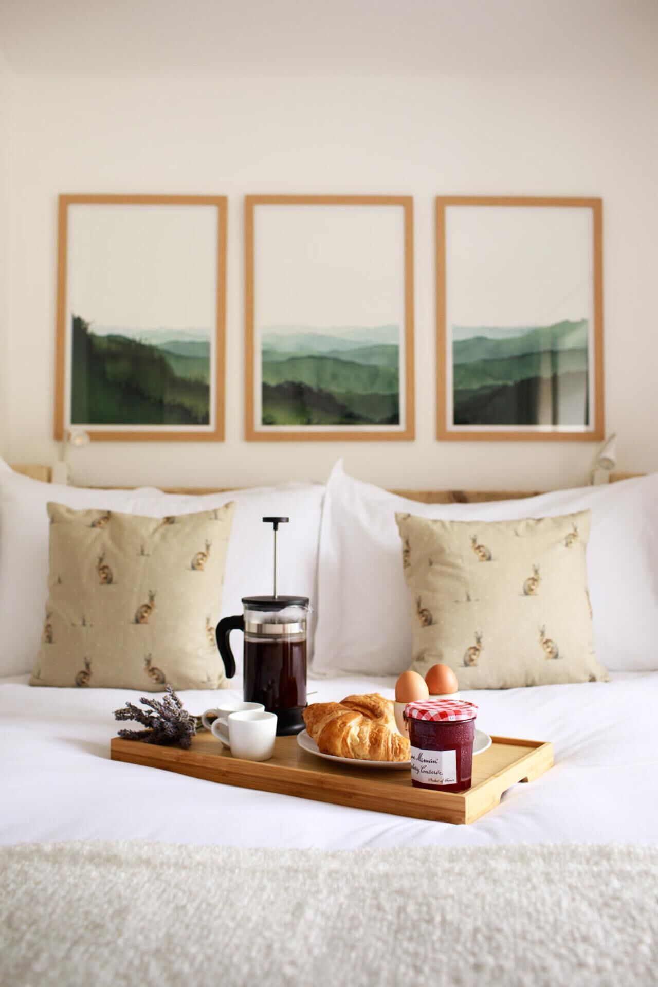 A neatly made bed with a tray of breakfast items including croissants, eggs, and jam, a coffee maker, and a cup of coffee.