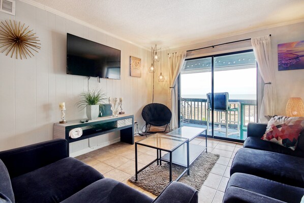 Relax in this stylish living room with a flat-screen TV, comfortable sofas, and a sliding glass door leading to a balcony with ocean views.
