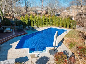 A rectangular swimming pool with a tiled deck, surrounded by a wooden deck with chairs and a table.