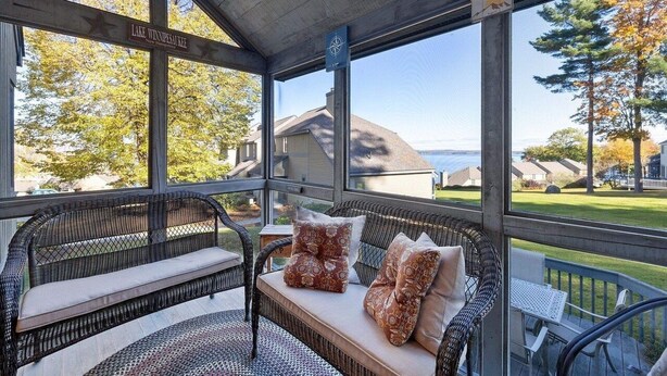 A covered outdoor seating area with wicker furniture and cushions, overlooking a residential neighborhood and a lake.