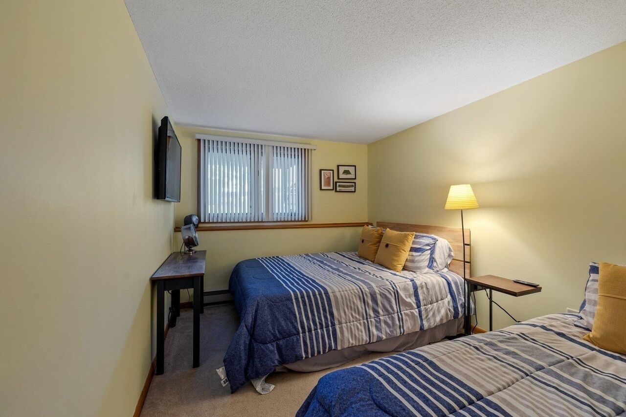 A hotel room with two beds, a desk, a lamp, and a window with blinds.