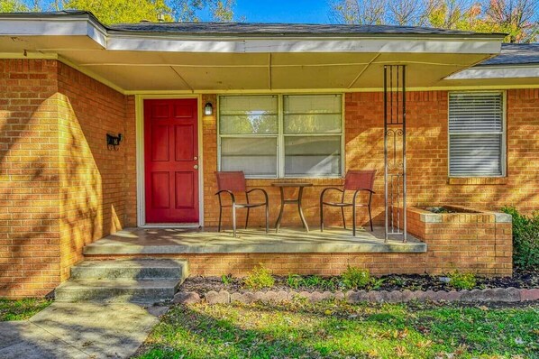 A brick house with a red door, two windows, and a small patio with two chairs.