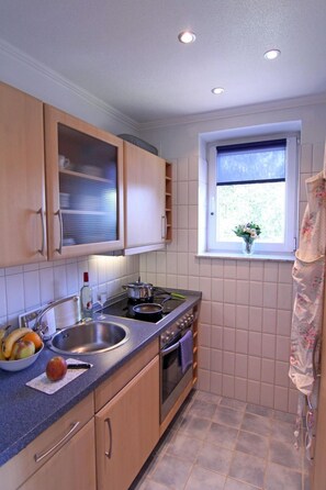 Een kleine keuken met houten kastjes, een roestvrijstalen spoelbak en een raam met uitzicht op groen.