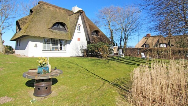 A thatched-roof house with a white exterior, a small garden with a flower pot, and a clear blue sky.